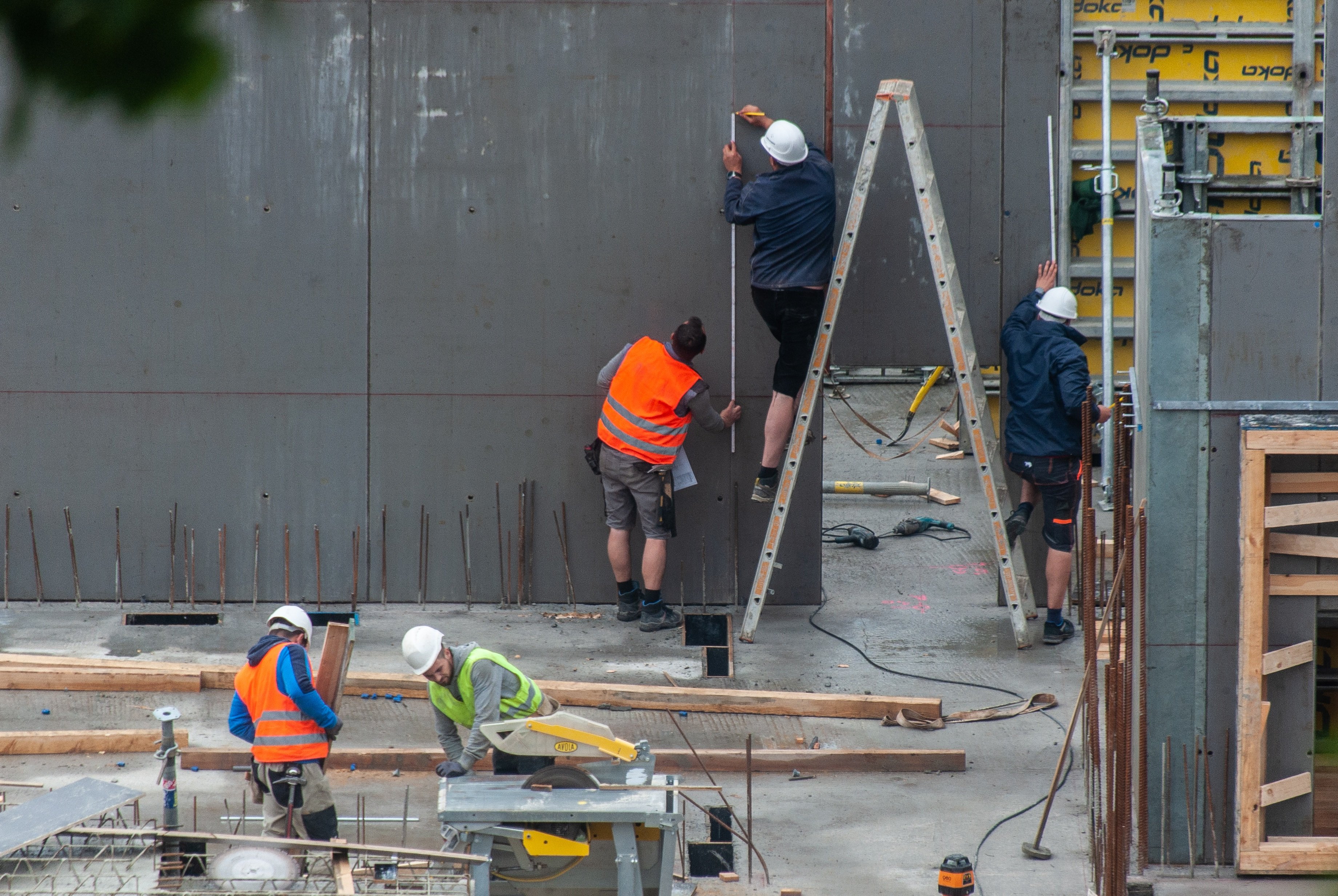Small crew working at construction site.  One worker stands on a ladder measuring and marking on the inside of concrete forms for a large column.