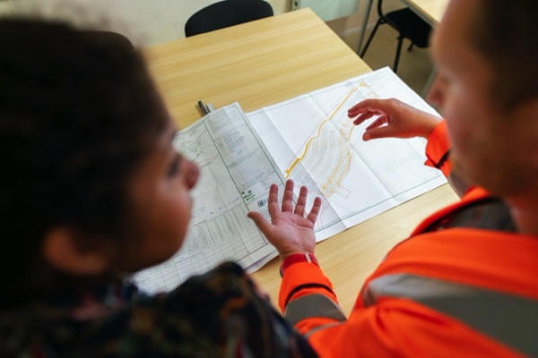 Looking over the shoulder of a person in construction orange suit standing next to person in business attire with construction plans displayed on table in front of them.
