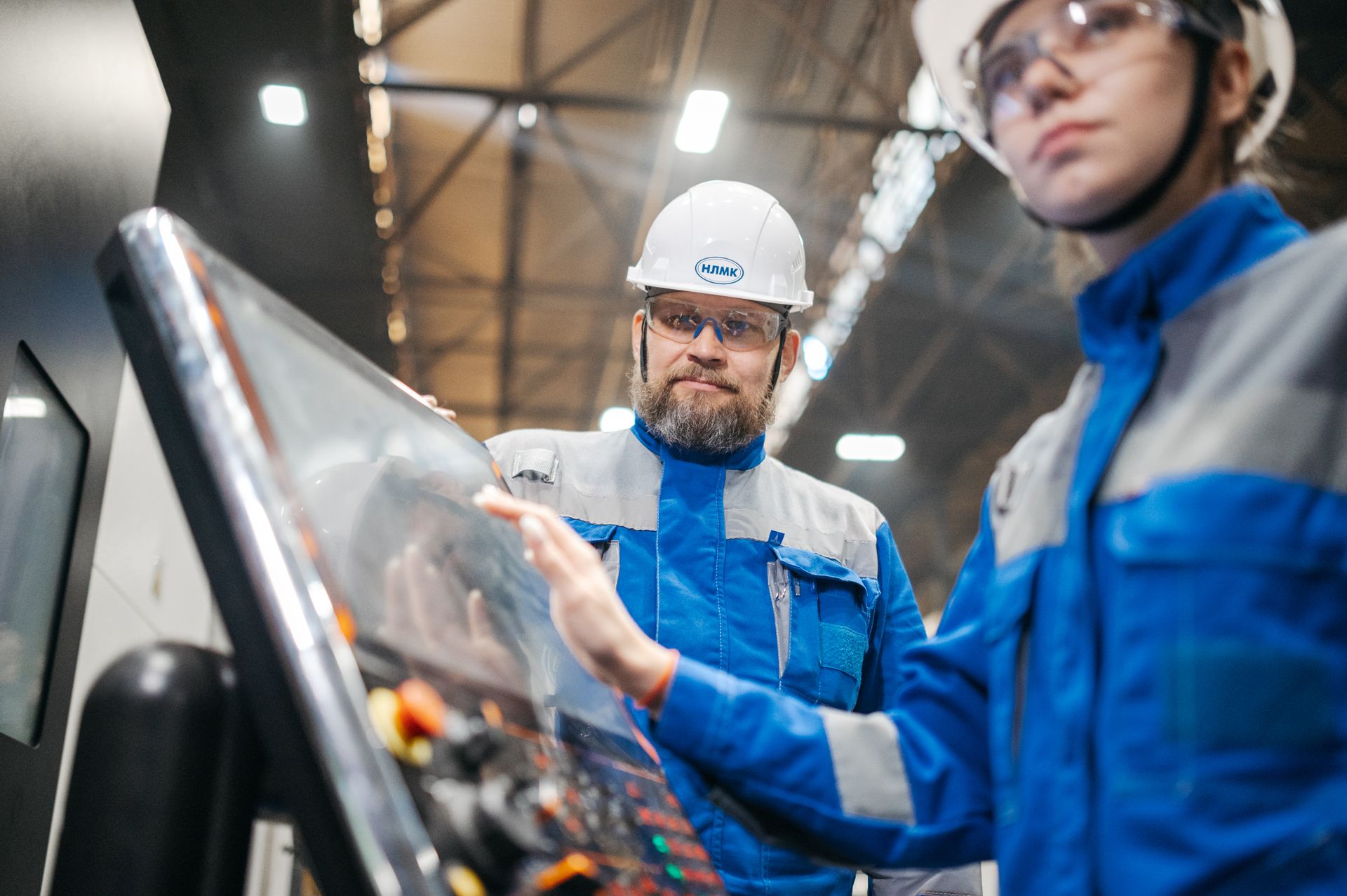 Two people dressed in industrial jumpsuits and PPE stand at a digital control panel. One is making adjustments via a touch screen on the panel.