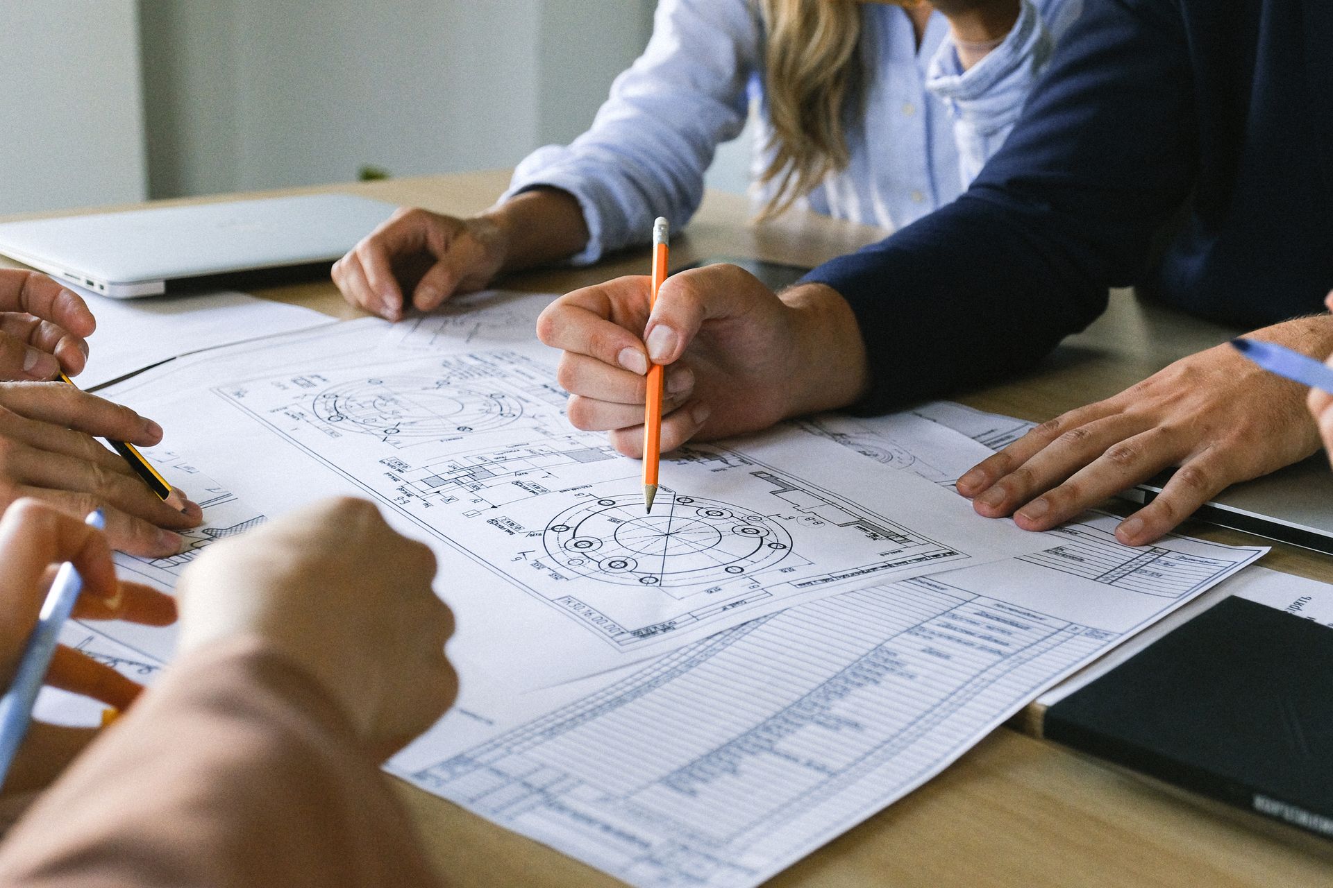 photograph showing medium shot of construction plans on desktop with 4 arms with hands extending into the shot, 2 with pens, making notes or pointing at the drawings.